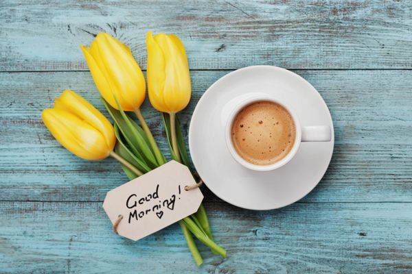 لیوان قهوه با گل های لاله زرد و یادداشت صبح بخیر روی میز آبی روستایی از بالا صبحانه در روز مادر یا روز زن