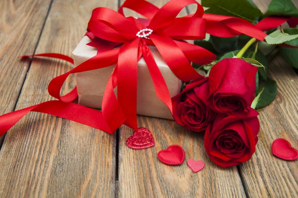 گل رز قرمز زیبا و جعبه هدیه با قلب در زمینه چوبی سفید