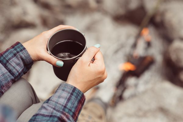 دست های مسافر زن که فنجان چای را در نزدیکی آتش در فضای باز نگه داشته است مفهوم ماجراجویی سفر گردشگری و کمپینگ کوهنورد در حال نوشیدن چای از لیوان در کمپ قهوه پخته شده روی آتش در طبیعت
