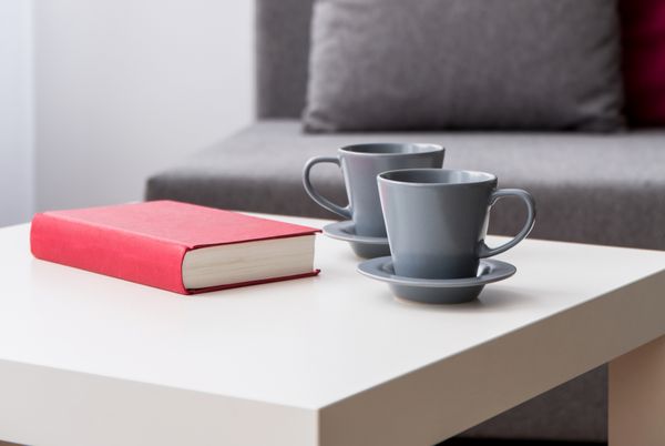 نمای نزدیک از یک میز قهوه با دو فنجان خاکستری و یک کتاب با جلد قرمز در کنار مبل خاکستری تار در پس زمینه