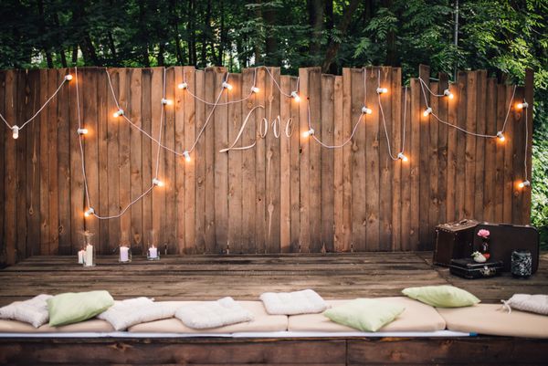دیوار چوبی تزئین شده با لامپ های برقی و علامت عشق چمدان ها و بالش های قدیمی روی زمین عروسی پذیرایی منطقه استراحت