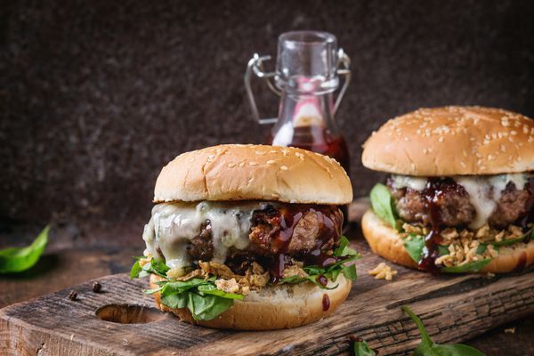 دو همبرگر با کتلت همبرگر گاو پیاز سرخ شده اسفناج سس سس کچاپ و پنیر آبی در نان های سنتی روی تخته خردکن چوب روی زمینه چوبی تیره سرو می شود
