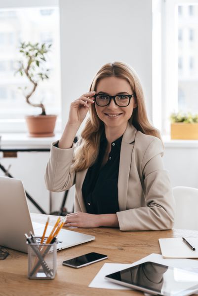 چگونه می توانم به شما کمک کنم؟ زن جوان زیبا با لباس هوشمند عینک خود را تنظیم می کند و به دوربین نگاه می کند در حالی که در اتاق کار خود در دفتر خلاق نشسته است