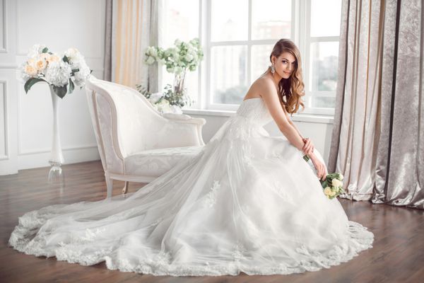 عروسی عروس با لباس زیبا روی مبل داخل خانه در فضای داخلی استودیو سفید مانند خانه نشسته است سبک عروسی مرسوم مد روز در طول کامل مدل قفقازی جوان جذاب مثل عروس