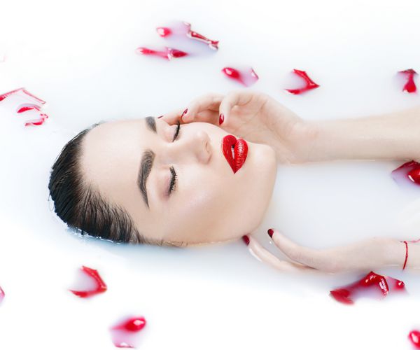 دختر مدل مد زیبا در حمام شیر لمس کردن پوست آبگرم و مفهوم مراقبت از پوست زن جوان زیبایی با گل رز قرمز در حال استراحت در حمام شیر ج و دست سالم جوانسازی