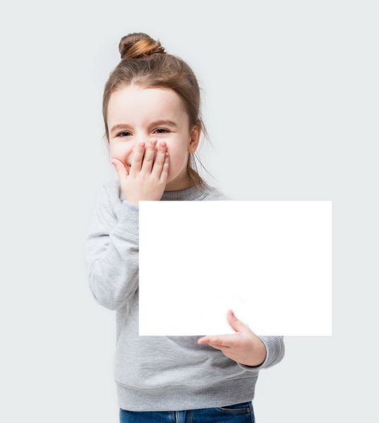 دختر بچه خوشحال می خندد با دست صورتش را می بندد دختر کوچولوی گیج که برای اطلاع شما پوستری در دست گرفته است پس زمینه خاکستری