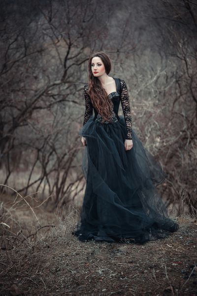 دختر زیبا با موهای بلند ایستاده در لباس مشکی ایستاده روی پس‌زمینه گوتیک جنگل‌های دمنده شاهزاده خانم جنگل هالووین بوهو تیره رنگ‌بندی مد روز رنگ خلاقانه