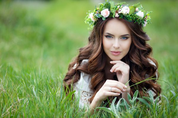 پرتره بهاری زن زیبا در تاج گل موهای فرفری بلند قرمز چشمان خاکستری لبخندی زیبا با لباس تابستانی سفید دراز کشیده روی چمن سبز نرم در فضای باز در بهار