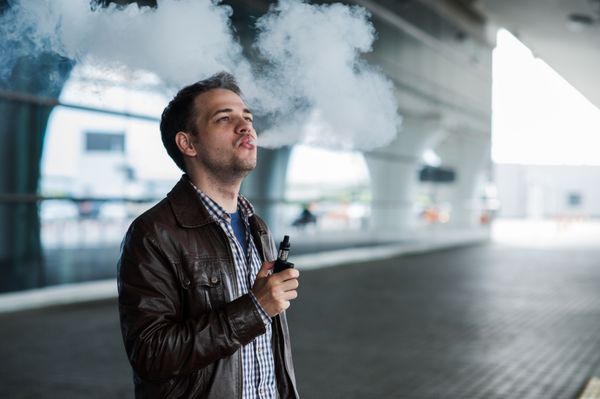 مرد جوان مسافر در حال کشیدن سیگار الکترونیکی در فضای باز نزدیک ترمینال فرودگاه