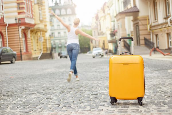 چمدان نارنجی در جاده در شهر و زن جوان در پس زمینه مفهوم تعطیلات تابستانی و سفر