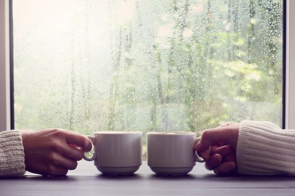 قهوه استراحت راحت برای دو نفر دو فنجان سفید در دست در پس زمینه ای از پنجره مرطوب با قطرات باران
