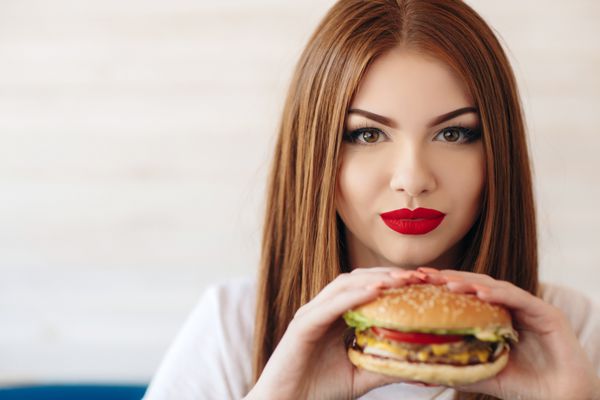 زن زیبا با موهای صاف و بلند قرمز آرایش روشن چشمان قهوه‌ای رژ لب قرمز مژه‌های بلند لاک صورتی در دست که یک همبرگر بزرگ در دست دارد پشت میز یک کافه نشسته تنها شام