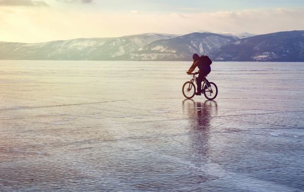 دوچرخه سوار تنها با کوله پشتی روی دوچرخه روی یخ دریاچه بایکال در پس زمینه آسمان غروب خورشید سطح یخ مفهوم ورزش زمستانی دوچرخه سواري