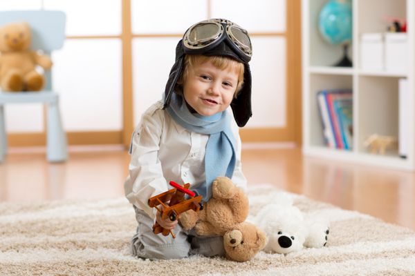 پسر بچه شاد در حال بازی با هواپیمای چوبی اسباب بازی و خرس عروسکی در اتاق کودکان