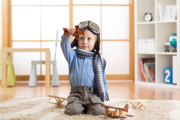 پسر بچه خوشحال در حال بازی با هواپیماهای اسباب بازی چوبی روی زمین در اتاق مهد کودک