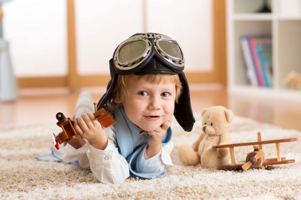 کودک خلبان یا هوانوردی که در خانه در اتاق مهد کودک با هواپیمای اسباب بازی بازی می کند مفهوم رویاها و سفرها