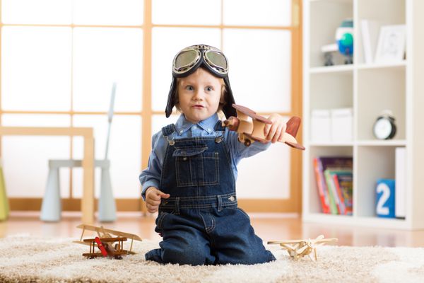 پسر بچه ای که کلاه ایمنی به سر داشت با هواپیمای اسباب بازی چوبی در اتاق بچه هایش بازی می کند