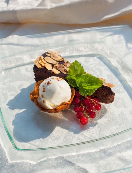 کیک بادام شکلاتی با فلفل دلمه ای شیرین بستنی وانیلی و توت قرمز روی بشقاب شیشه ای و رومیزی سفید
