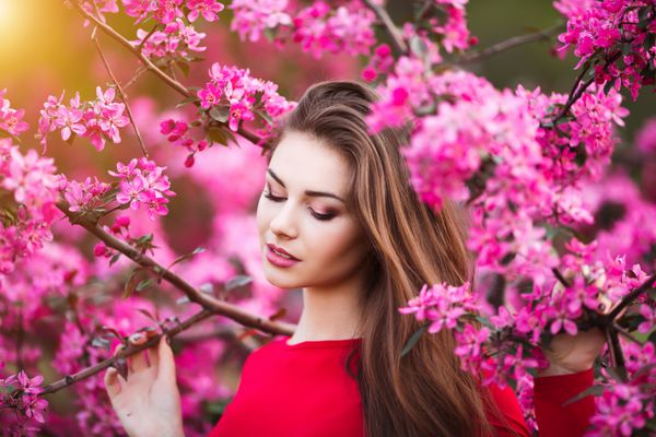 لمس بهاری زن جوان و زیبای شاد با لباس قرمز از گل های صورتی تازه و نور خورشید در پارک شکوفه در غروب آفتاب لذت می برد
