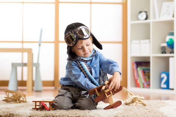 پسر بچه ای که کلاه ایمنی به سر داشت در اتاق بچه هایش با هواپیماهای اسباب بازی چوبی بازی می کند