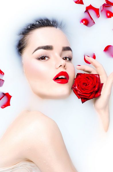 دختر مدل جوان زیبا با آرایش روشن و گل رز قرمز در حال استراحت در حمام شیر