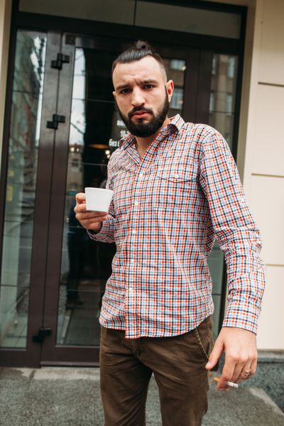 مرد جوانی که دارای فضای آزاد دود است هیپستر شیک در حال استراحت از محل کار با یک فنجان قهوه و سیگار مفهوم اعتیاد بد عادت سلامتی آرامش