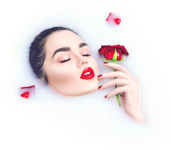 دختر مدل زیبا با آرایش روشن که گل رز قرمز را در دست گرفته و در حمام شیر استراحت می کند