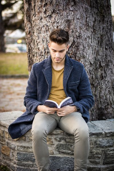دانش آموز پسر در فضای باز کتاب می خواند مرد جوان خوش تیپ بیرون روی نیمکتی در پارک شهر نشسته کتاب می خواند