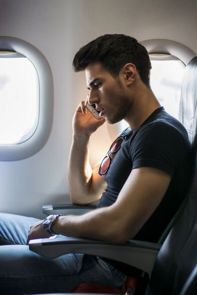 نمای جانبی مرد جوان خوش تیپ در مقابل پنجره هواپیما که نشسته و با تلفن صحبت می کند