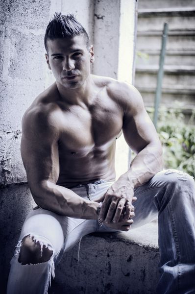 مرد جوان لاتین عضلانی بدون پیراهن با شلوار جین روی دیوار بتونی نشسته و به دوربین نگاه می کند