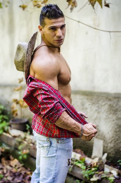 مرد جوان خوش‌تیپ و عضلانی که پیراهن را از تن در می‌آورد شلوار جین و کلاه حصیری بر سر دارد در فضای باز