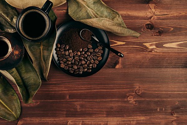قهوه داغ در فنجان سیاه با دانه‌ها برگ‌های خشک و گلدان ترکی با فضای کپی روی پس‌زمینه تخته چوبی قدیمی قهوه‌ای نمای بالا سبک روستیک