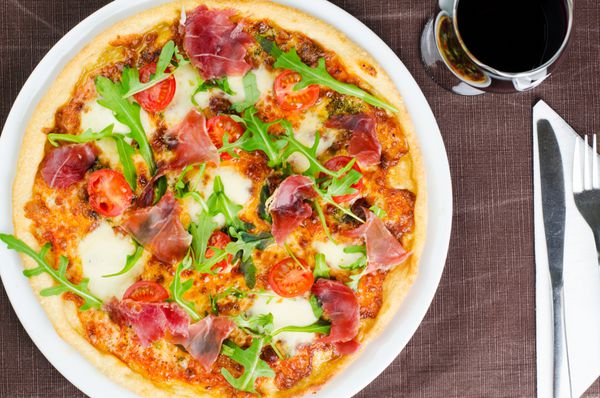 پیتزای خوشمزه ایتالیایی با پروشوتو گوجه فرنگی تازه و روکولا در کنار یک لیوان شراب قرمز