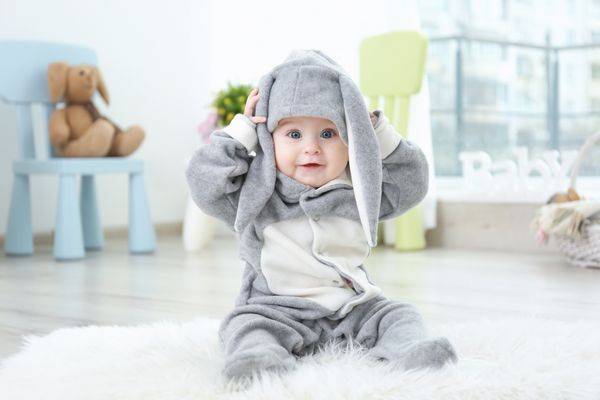 نوزاد کوچولوی ناز با لباس اسم حیوان دست اموز که روی فرش خزدار در خانه نشسته است