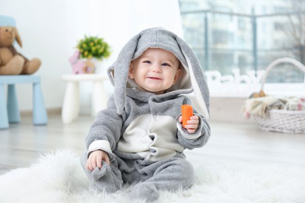 نوزاد کوچولوی ناز با لباس خرگوش که روی فرش پشمالو نشسته و هویج می‌خورد