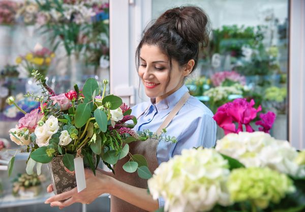 گلفروش زن زیبا که گلدانی با گل در گلفروشی در دست دارد