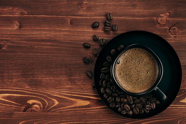 قهوه داغ در فنجان سیاه با خامه و دانه‌ها با فضای کپی روی پس‌زمینه تخته چوبی قدیمی قهوه‌ای نمای بالا سبک روستیک