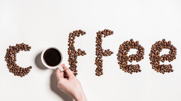 نمای بالای قهوه کلمه ساخته شده از دانه های قهوه و دست انسان که فنجان قهوه سیاه را در دست دارد