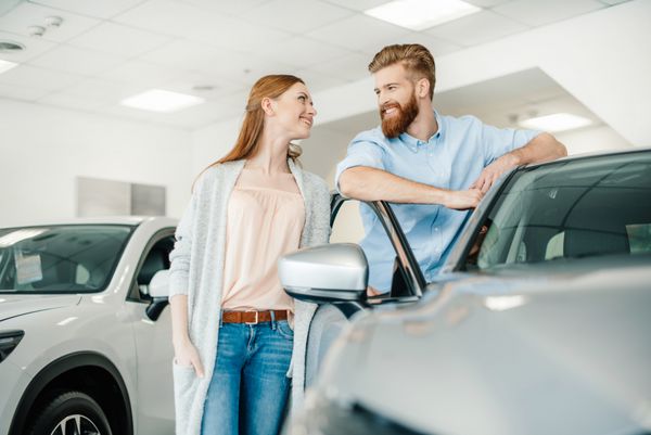 زوج جوان شادی که نزدیک ماشین جدید ایستاده اند و به یکدیگر لبخند می زنند