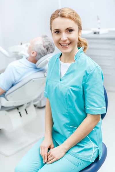 دندانپزشک حرفه ای با لباس پزشکی در کلینیک نشسته و در مقابل دوربین لبخند می زند