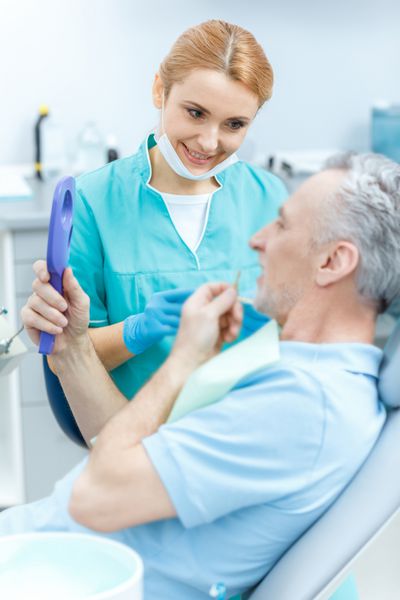 دندانپزشک حرفه ای خندان به بیمار بالغ که آینه در دست دارد نگاه می کند