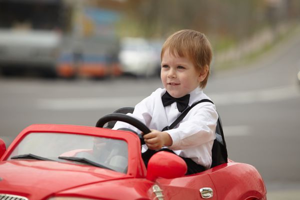 پسر بچه ای با پیراهن سفید در ماشین اسباب بازی قرمز در خیابان