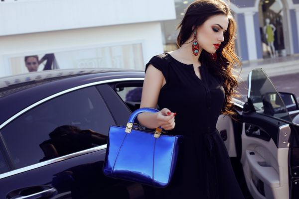 زنی با لباس زیبا با کیف در کنار یک خودروی مجلل ژست گرفته است