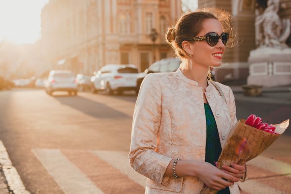 زن زیبای جوان و شیک در حال قدم زدن در خیابان شهر در غروب آفتاب تعطیلات اروپا دسته گل لاله در دست دارد گل ترند بهاری عینک آفتابی خندان لوکس لب قرمز لباس مد روز ژاکت