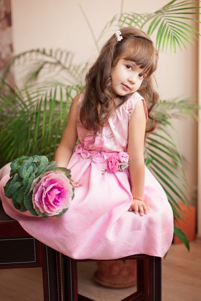 نمای مدل دختر کوچولوی زیبا با لباس صورتی لوکس و ژست گل در خانه روی میز