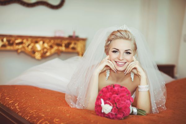 عروس زیبا با لباس عروس در فضای داخلی مجلل با جواهرات الماس که در خانه ژست گرفته و منتظر داماد است دختر شاد پولدار عاشقانه با لباس عروس خندان آماده نهایی برای عروسی است