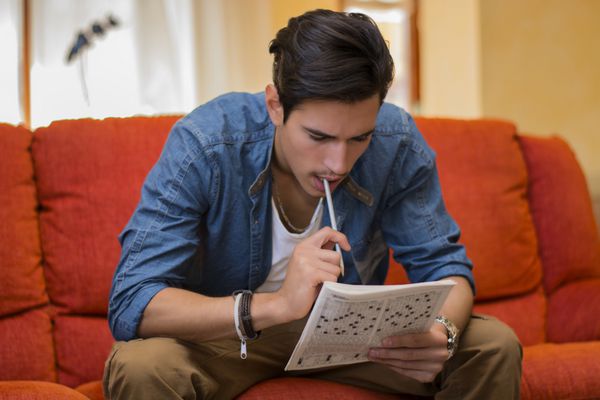 مرد جوانی نشسته و جدول کلمات متقاطع را انجام می دهد و با مداد به دهانش متفکرانه به مجله نگاه می کند در حالی که سعی می کند به جواب سرنخ فکر کند