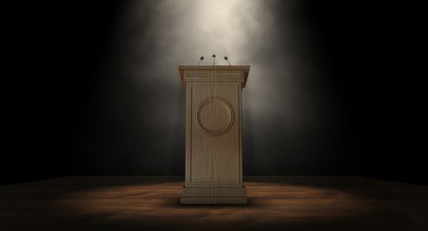 یک تریبون سخنرانی چوبی با سه میکروفون کوچک که روی پس‌زمینه‌ای تیره با یک نورافکن متصل شده‌اند