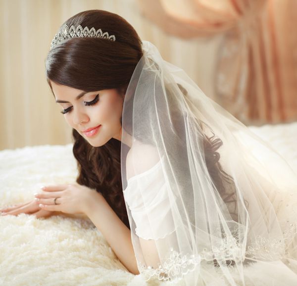 پرتره عروس زیبا با حجاب مد که در صبح عروسی روی تخت ژست گرفته است آرایش دختری با مدل موی موج دار بلند لباس عروسی