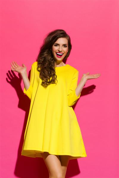 شادی در برابر دیوار صورتی دختر زیبای خندان با لباس مینی زرد که با دستان دراز ژست گرفته است استودیو سه چهارم روی پس‌زمینه صورتی عکس گرفته شده است